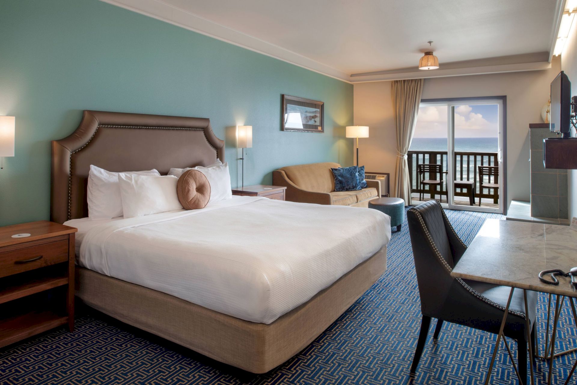 A seaside hotel room with a large bed, sofa, lamps, and a balcony view of the ocean under sunny skies.