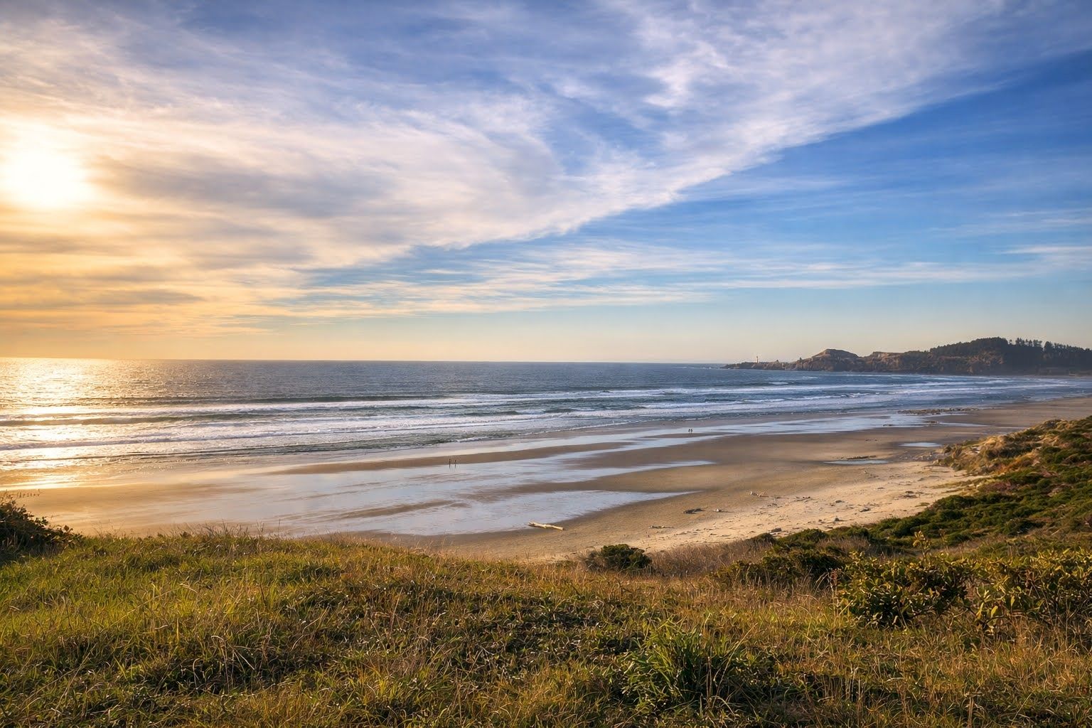 A wide sandy beach with ocean waves at sunrise, golden light on the shore, and grassy dunes in the foreground.