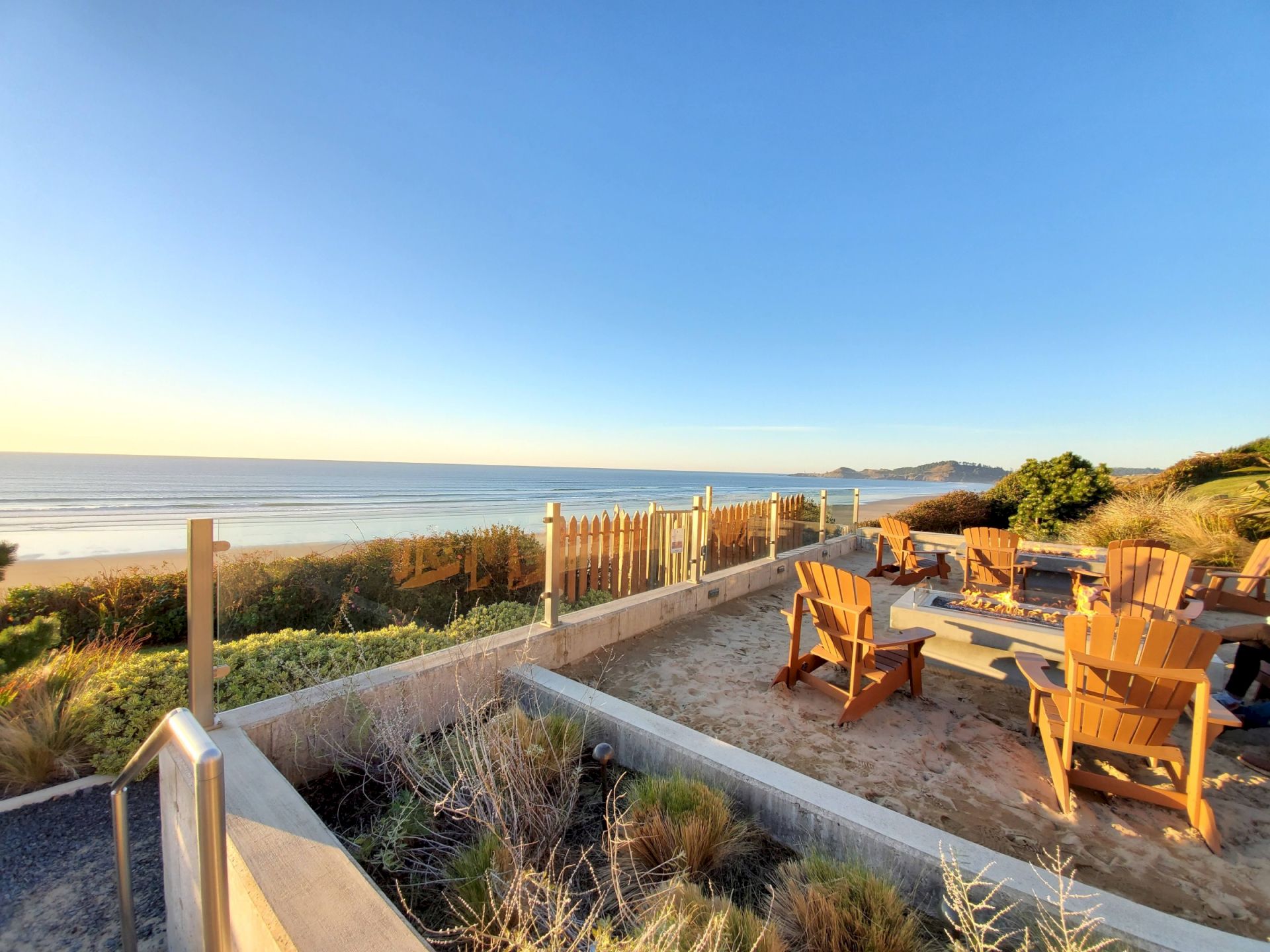 A beachfront deck with wooden chairs, a firepit, dunes and an ocean horizon at sunset.