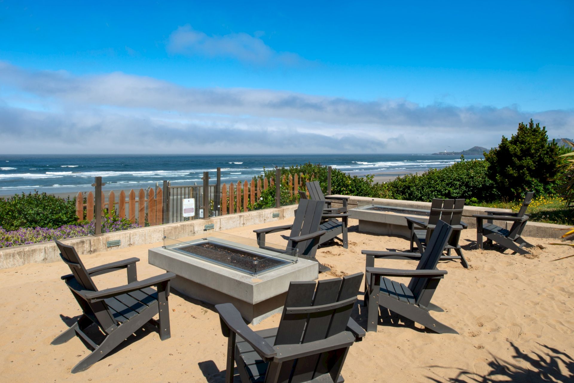 Beachside patio with wooden chairs around a fire pit, overlooking waves and a cloudy blue sky.