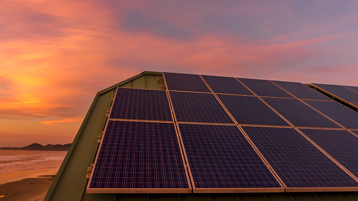 Solar panels line a rooftop at sunset, glowing orange skies over a calm shoreline.