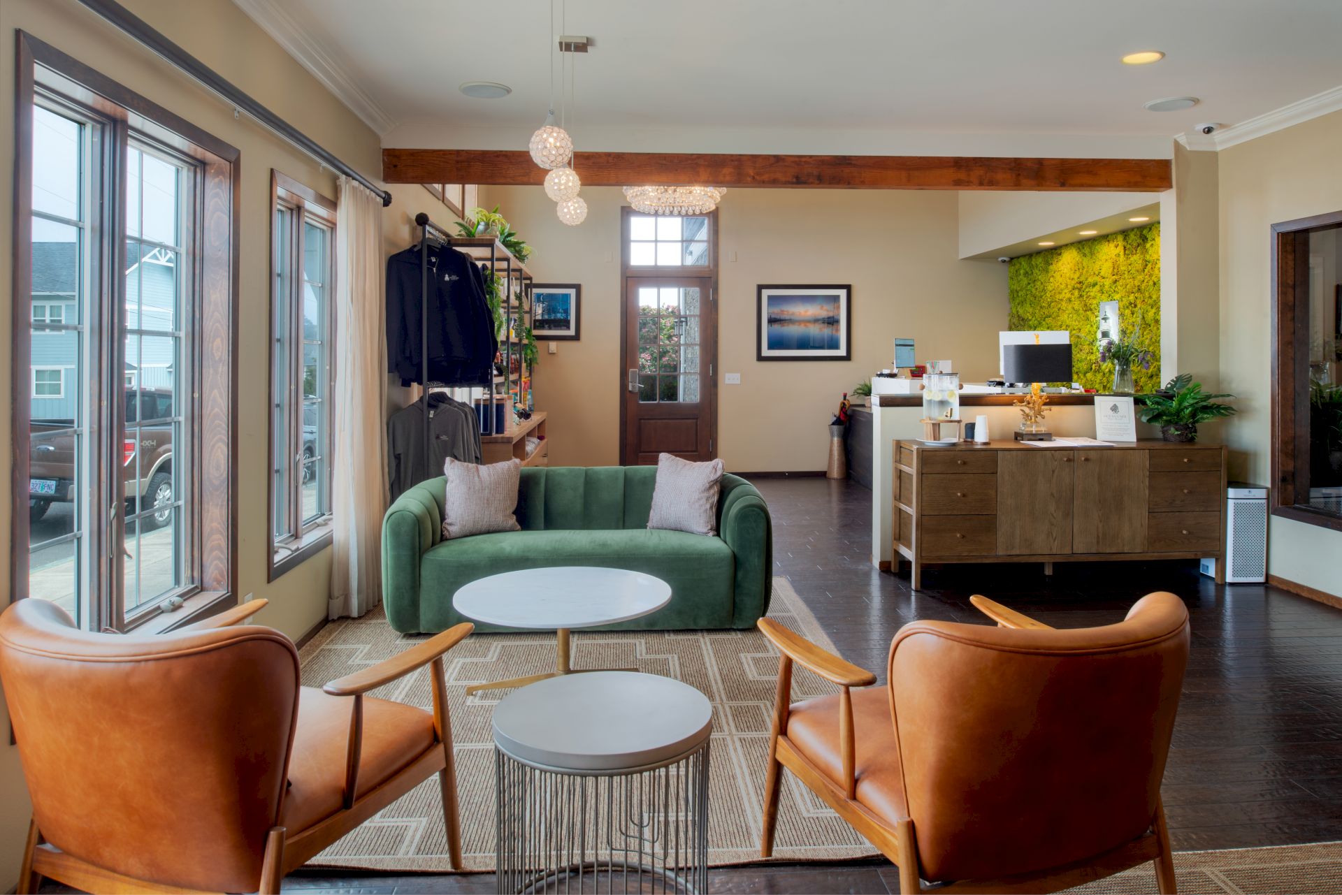 A modern, cozy showroom or lobby with a green sofa, two tan leather chairs, white round tables, and a small metal side table, near large windows and a reception desk.