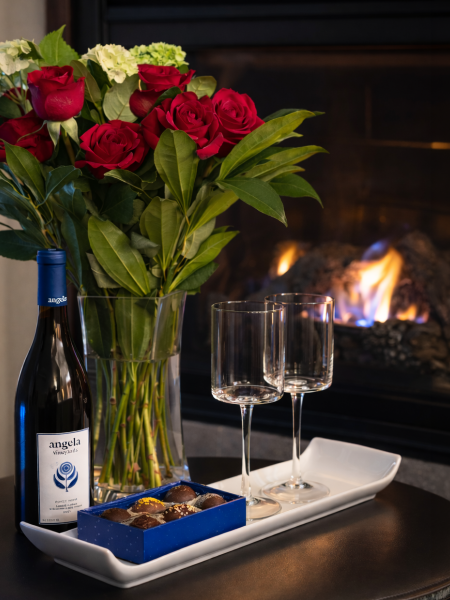 A tray with a bottle of wine, two glasses, a bouquet of red roses, and chocolates beside a crackling fireplace.