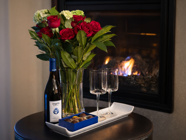 A tray with a bottle of wine, two glasses, a bouquet of red roses, and chocolates beside a crackling fireplace.