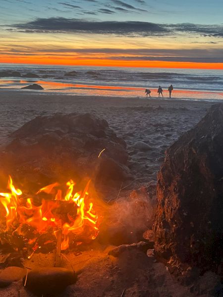 A beach bonfire glows beside rocks at sunset, silhouettes of people near the water as the sky fades to orange and blue.