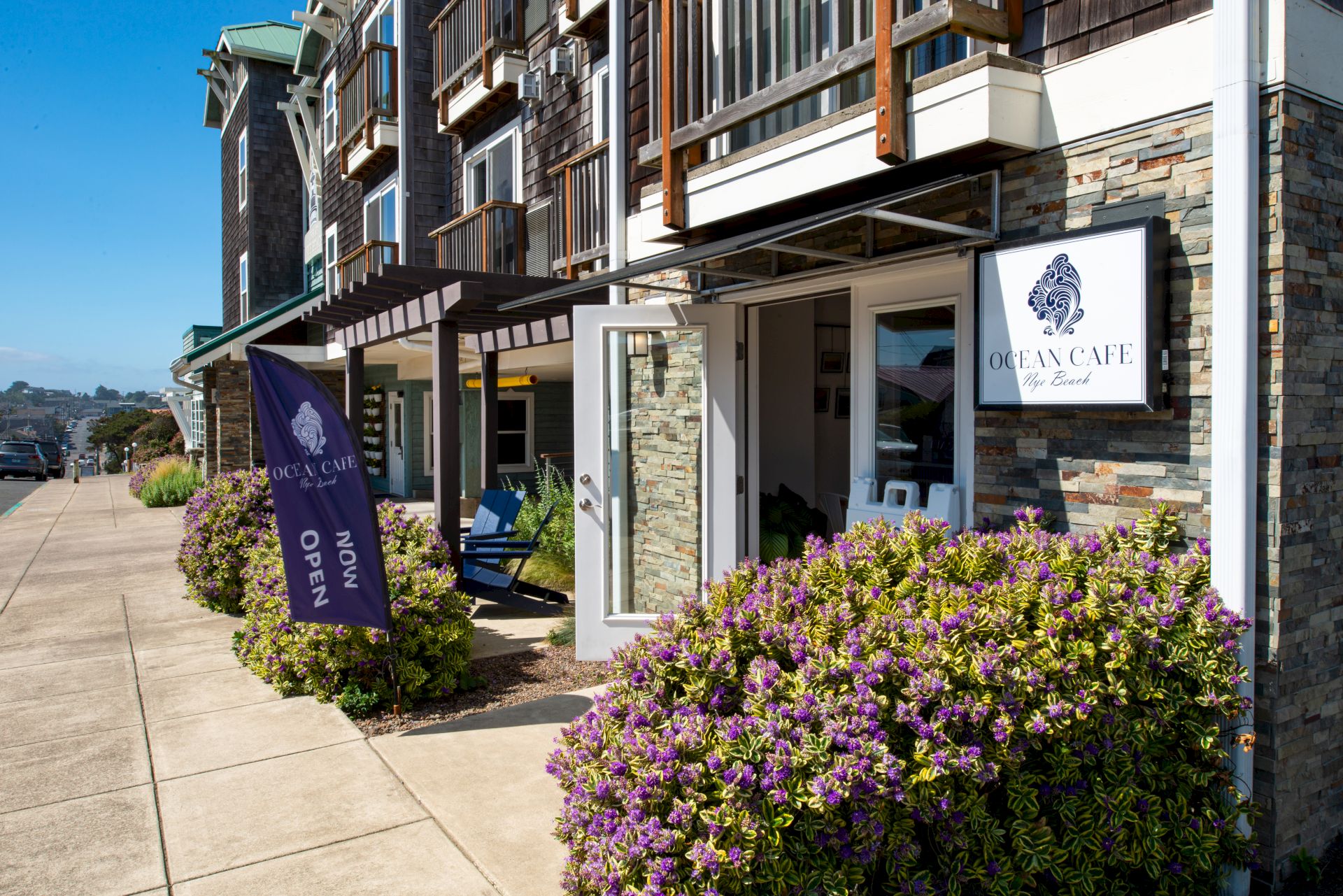 A street view of a modern building with stone siding, hanging awnings, a sign for "GlenCaine" or similar, purple flowering shrubs, and a sidewalk.