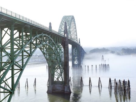 A large green steel bridge extends over a misty river, with arches fading into fog, and wooden pilings sticking up from the calm water.