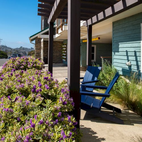 A sunny coastal scene with a wooden walkway, sandy beach, flowering plants, and a person sitting on a bench under a shaded awning, sea visible.