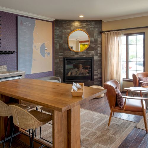 A sunlit rustic living room with a wooden coffee table, TV on the wall, stone fireplace, leather chair, and large windows showing outdoors.