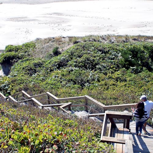 Staircase winds down a grassy, dune-covered hillside to a sandy beach; two people admire the view at a wooden overlook.