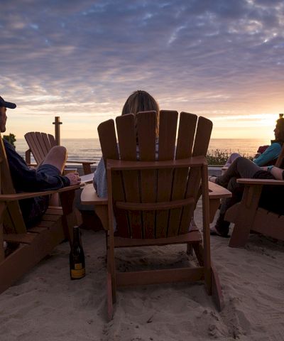 A group of people sits in wooden beach chairs around a small fire at sunset on the sand, enjoying drinks and conversation by the ocean.