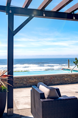 A sunny beachfront patio with a pergola, wicker furniture, a stone planter, and an ocean view with waves beyond a low wall.