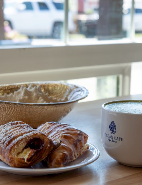 A coffee cup and a plate with croissants beside a bowl on a wooden table near a window.