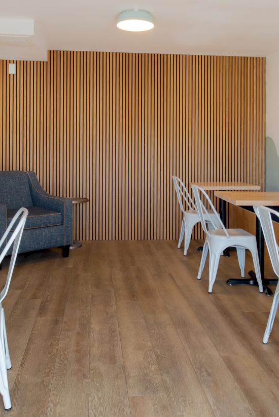Small modern cafe with light wood floors, white chairs, wooden tables, a blue-gray sofa, and a carved wooden slat wall at the back; soft wave mural on right wall.