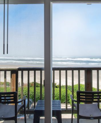 A seaside balcony with two chairs, curtains, and a wide ocean view with waves under a cloudy sky.
