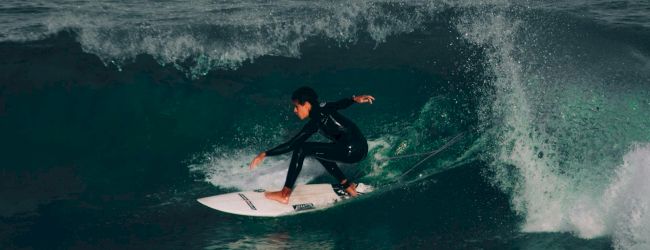 A surfer rides a small wave, crouched low on a white board, carving through teal water as a spray curtain erupts behind.