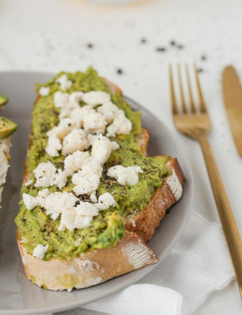 Slices of crusty bread topped with green pea or herb spread, crumbled white cheese, and a neatly plated dish with gold cutlery nearby.