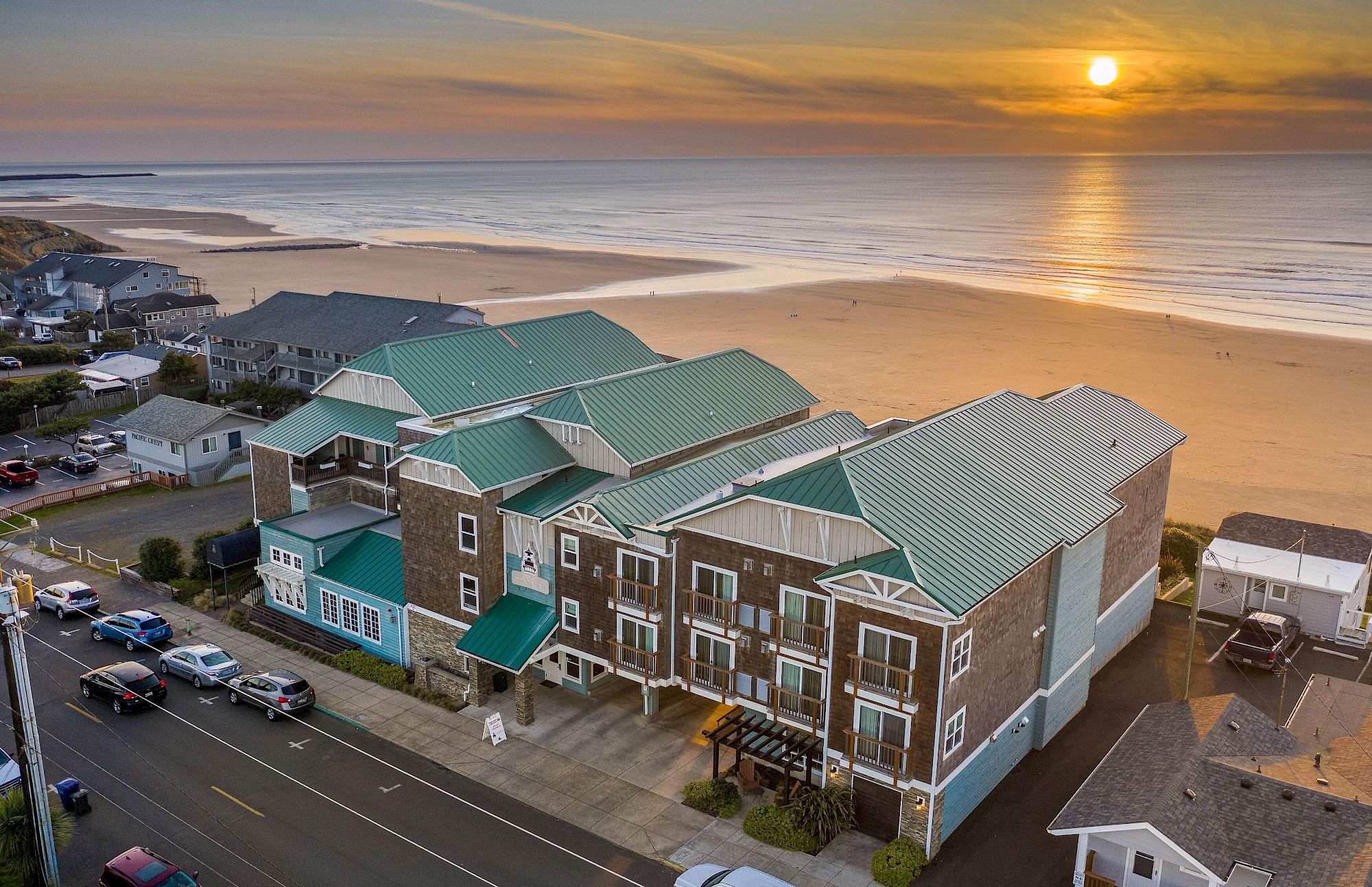 Aerial view of a beachfront hotel at sunrise with green roofs, parked cars, and a wide sandy shoreline where the sun reflects on calm waves.