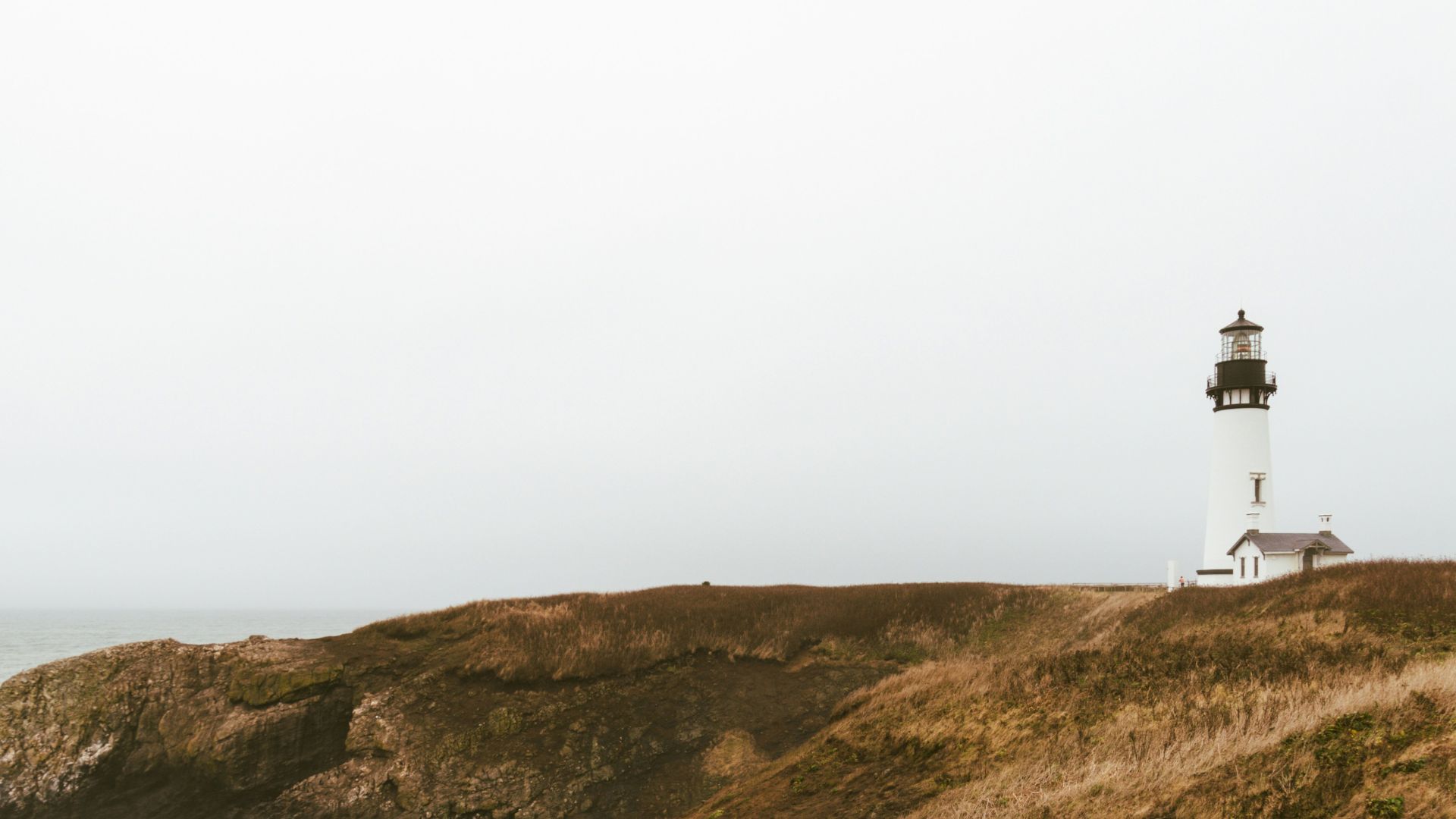 A tall white lighthouse stands on a grassy cliff by the ocean under a pale sky.