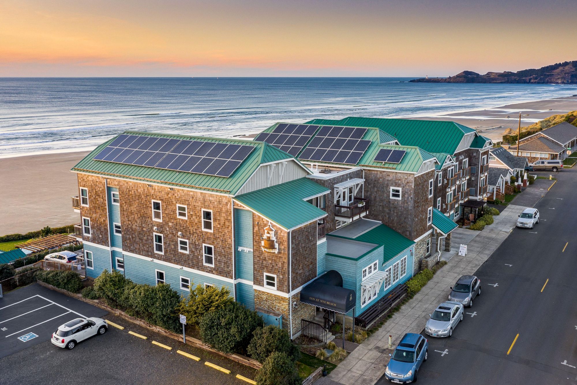 A coastal building complex with solar panels on green roofs, brick walls, blue accents, and cars parked along a seaside street at sunset.