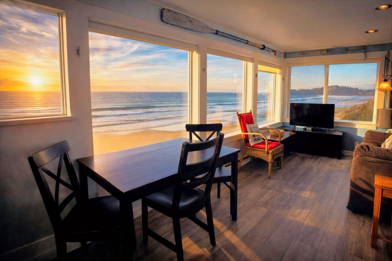 A cozy seaside living room with dining table, chairs, TV, and large ocean-view windows at sunset.