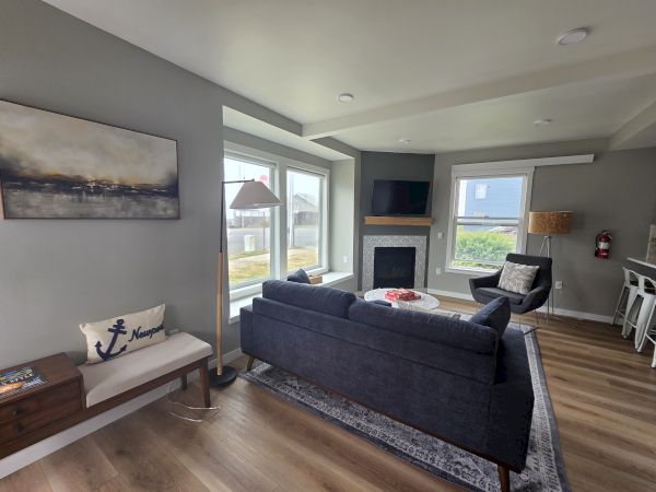 A modern living room with a dark sofa, TV over a fireplace, large windows, and an upholstered armchair.