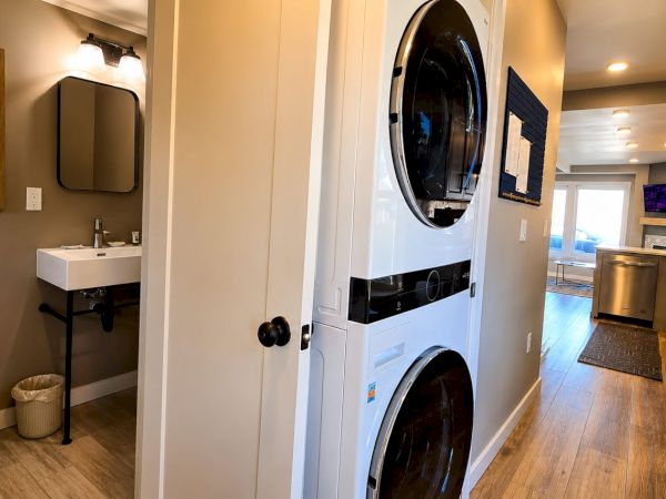 A modern laundry nook with a washer/dryer, small sink, and an open hallway leading into a bright kitchen area.