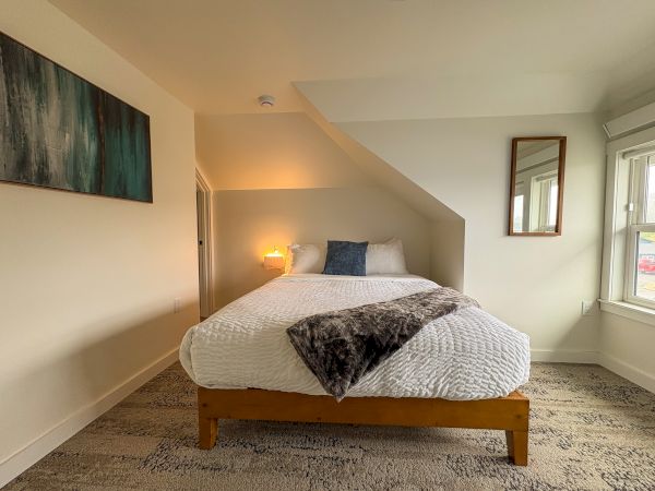 A cozy bedroom with a neatly made bed, pillows, bedside lamp, wall art, and windows along the walls, illuminated by warm light.