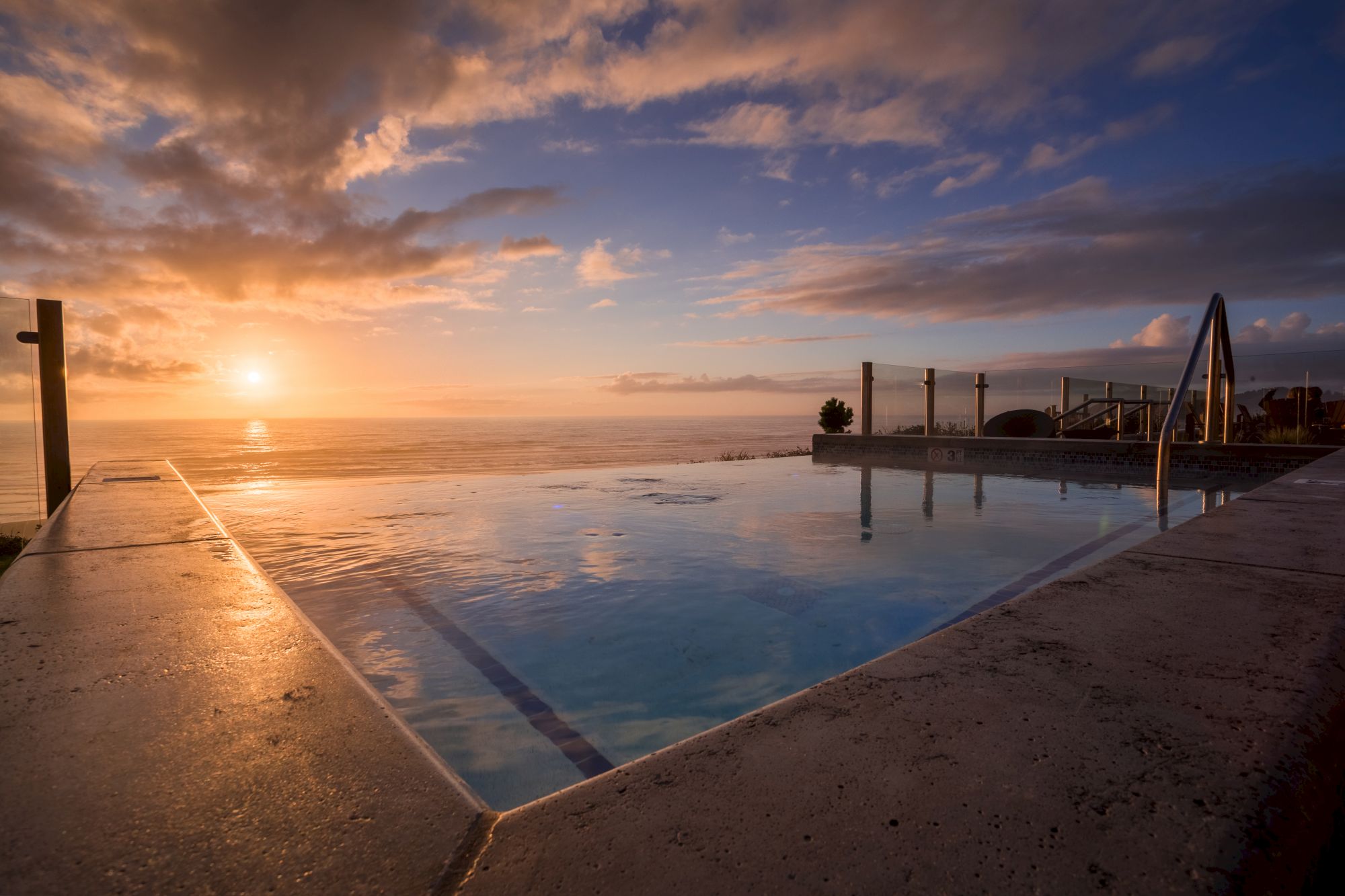 Sunset over the ocean beside a reflecting pool, with a cloudy orange sky and calm water.