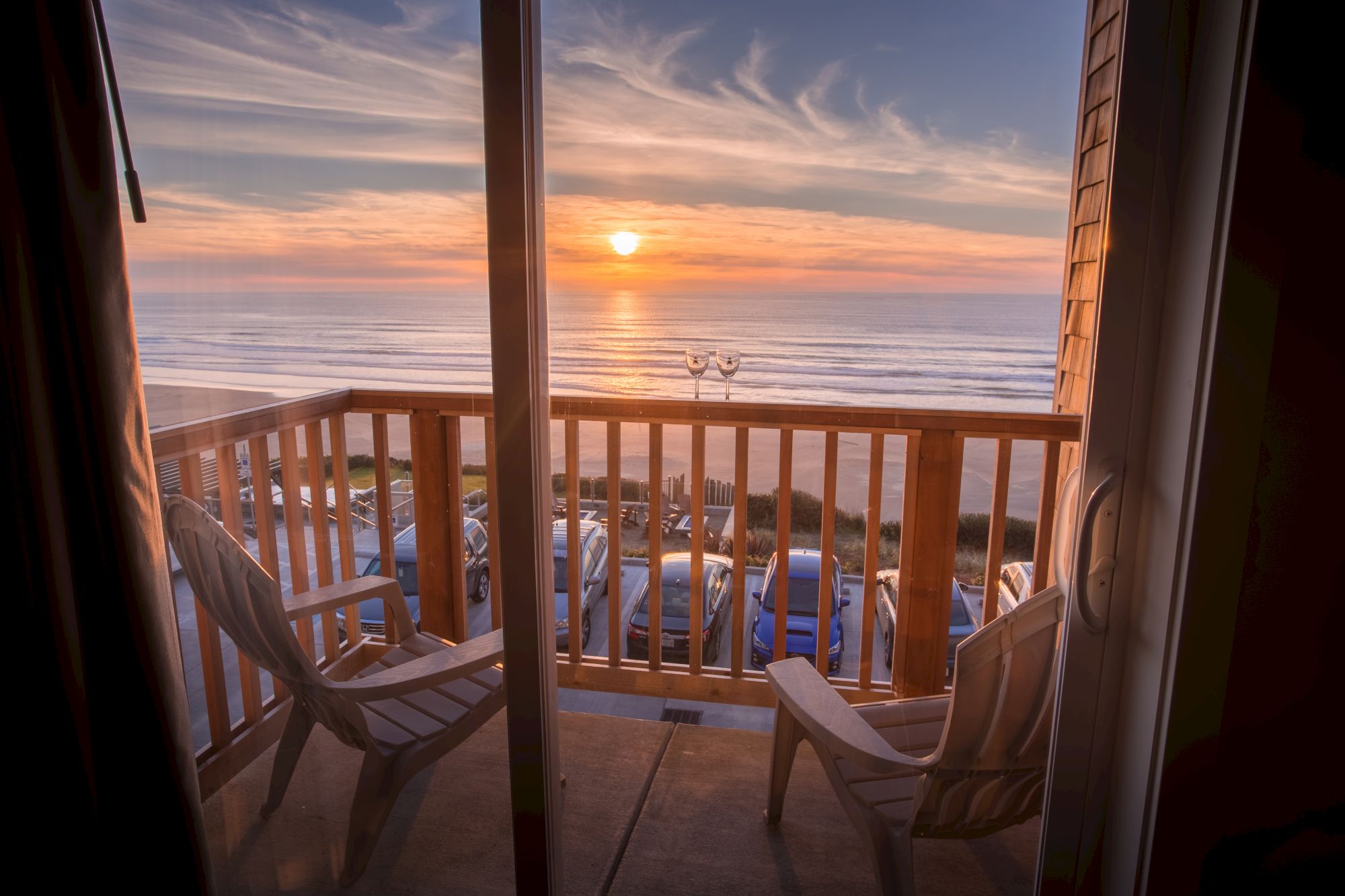 Balcony view at sunset: wooden railings, two empty wine glasses, chairs, parked cars, and the ocean glowing in the distance.