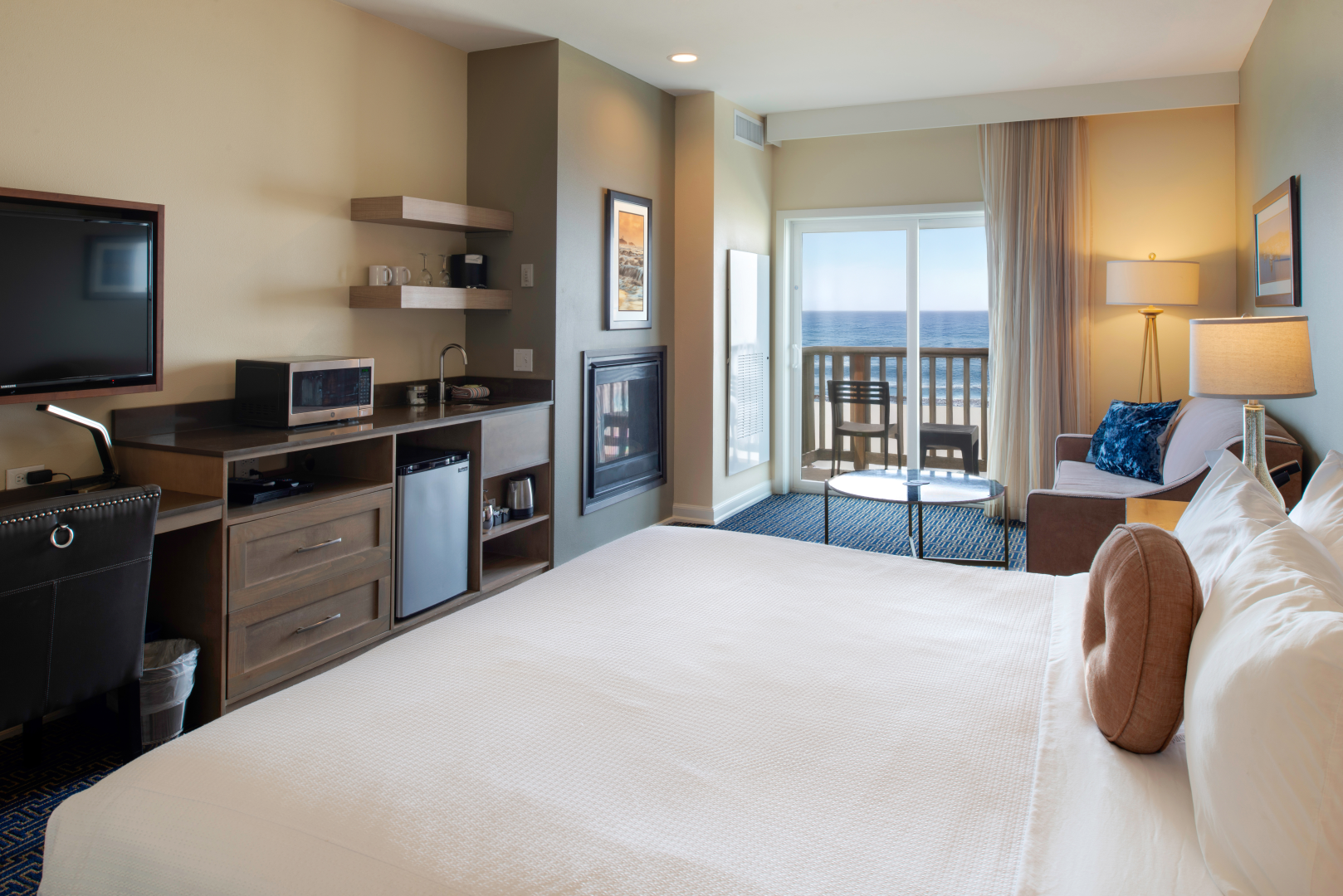 A modern hotel room with a large bed, small kitchenette, and ocean view through sliding doors, lit by warm lamps.