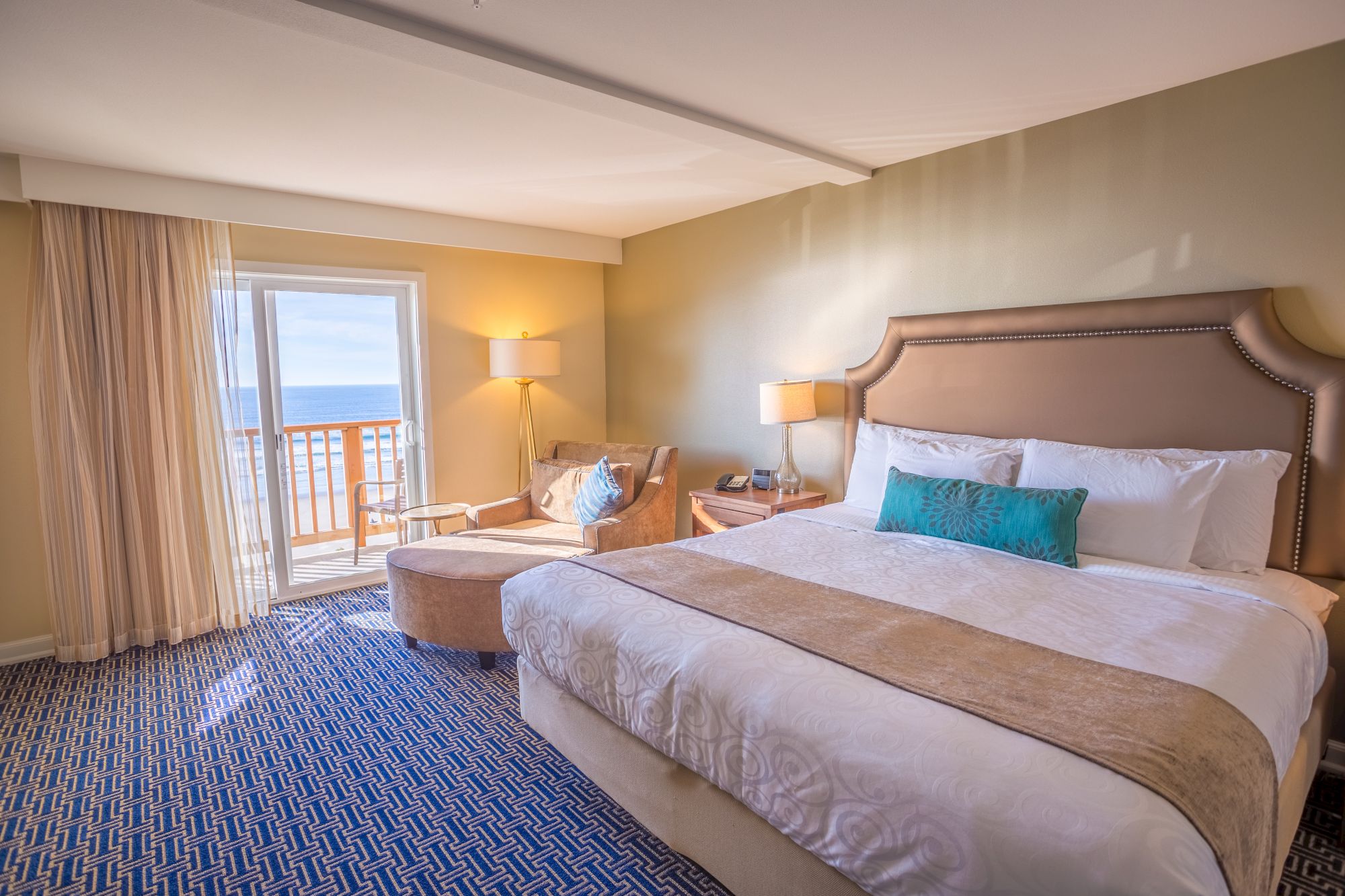 A bright hotel bedroom with a large bed, lamps, and ocean view through open balcony doors, with curtains and blue patterned carpet.